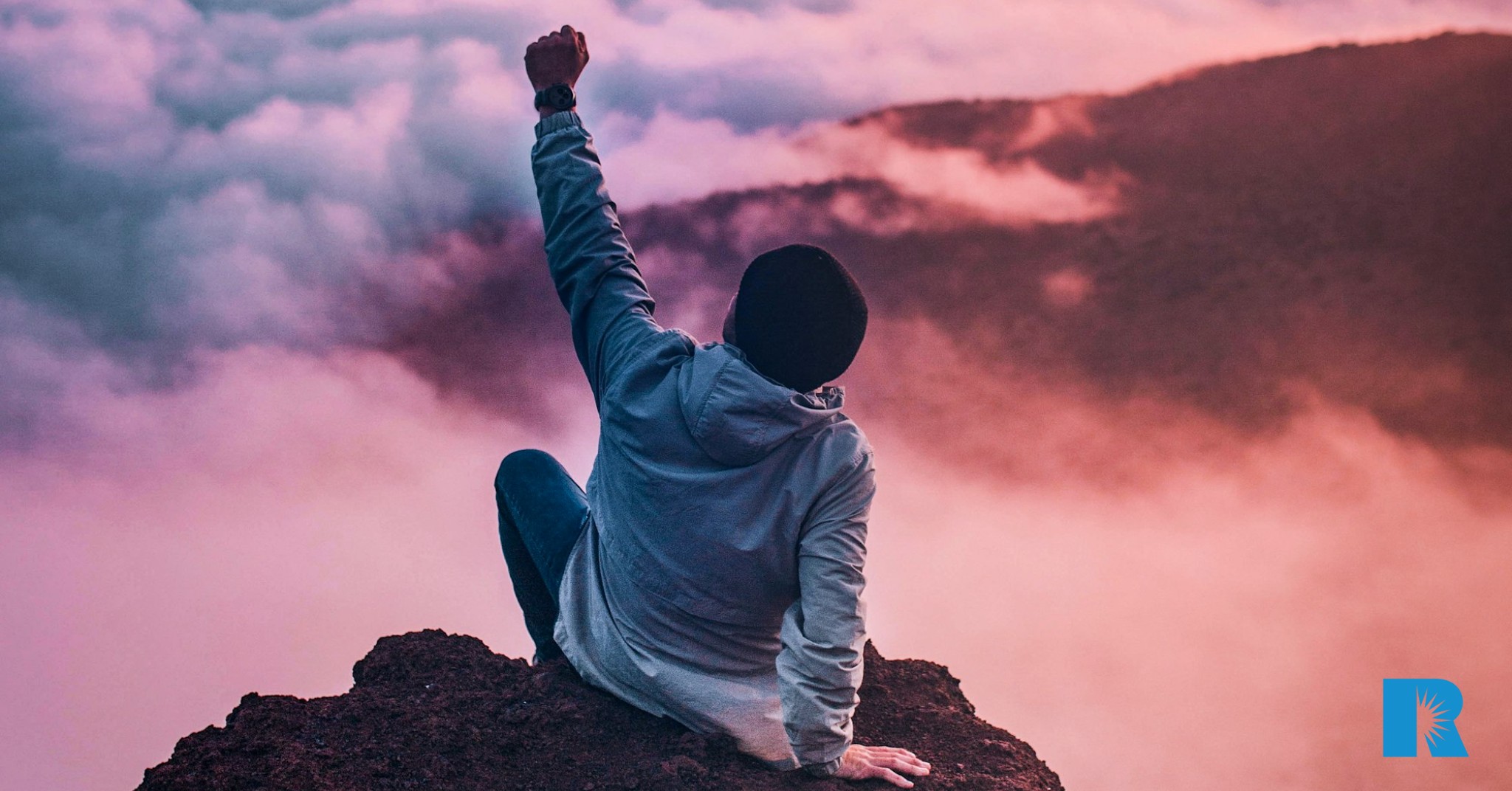 A man sits atop a mountaintop, raising his fist in defiance.